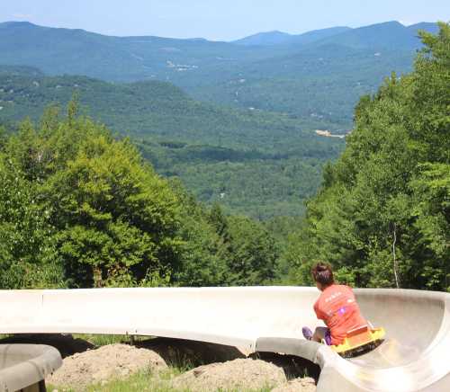 A child rides a summer slide down a hill, with lush green mountains in the background under a clear blue sky.