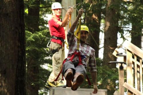 A man in a plaid shirt zip lines through a forest, while an instructor in a red shirt prepares behind him.