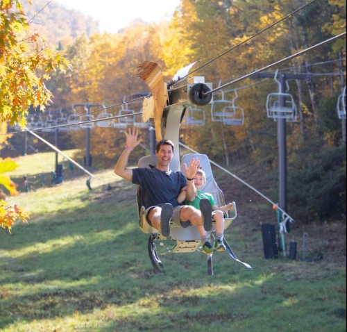 A man and a child happily ride a chairlift through a colorful autumn landscape.