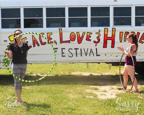 Two women hula hooping in front of a colorful bus with "Peace, Love & Hippies Festival" painted on it.