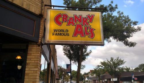 Sign for "Cranky Al's World Famous" hanging outside a brick building, with trees and a cloudy sky in the background.