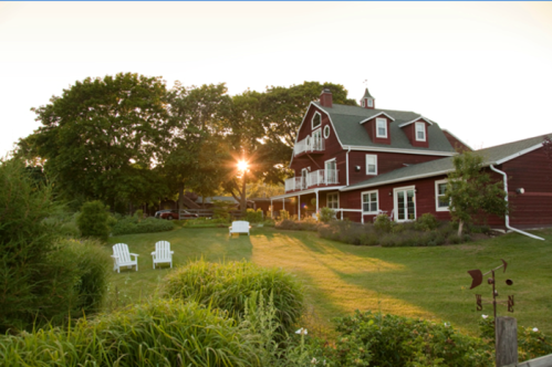 A red barn-style house surrounded by lush greenery, with a sunset shining through the trees in the background.