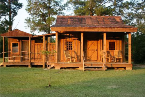 A rustic wooden cabin with a metal roof, featuring a porch and rocking chairs, surrounded by greenery.