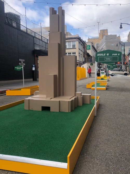 A large, abstract brown sculpture stands on a green turf area, part of a mini-golf course in an urban setting.