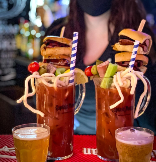 Two large cocktails topped with mini burgers and garnished with vegetables, alongside three smaller beers.