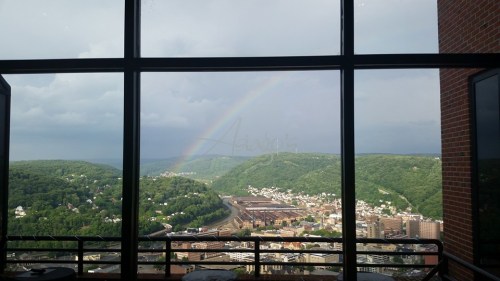 A view from a high-rise window showing a rainbow over green hills and a town below, with cloudy skies.