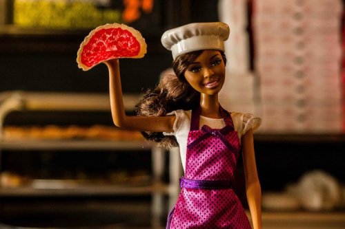 A doll wearing a chef's hat and apron holds a slice of pink cake, standing in a bakery setting.