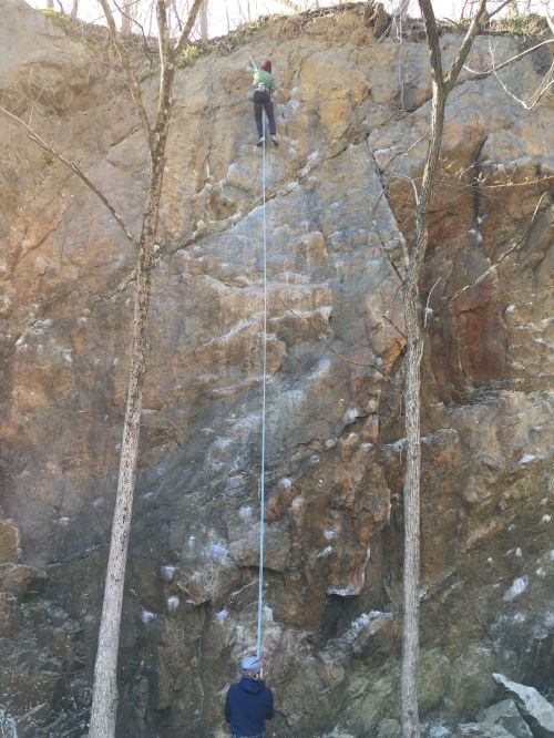 A person climbs a rocky cliff while another holds a safety rope below, surrounded by trees.