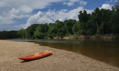 A bright orange kayak rests on a sandy riverbank, surrounded by lush green trees and a partly cloudy sky.