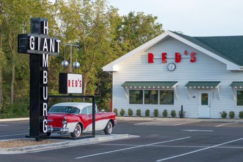 A vintage red and white car parked outside a diner named "Red's," featuring a large sign that reads "Giant Hamburg."