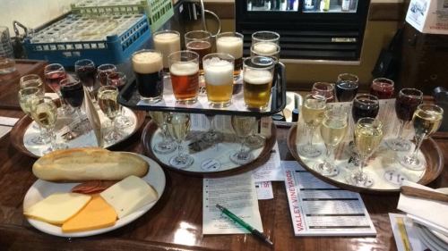 A wooden table with various glasses of beer, wine, and a platter of cheese, salami, and a breadstick.