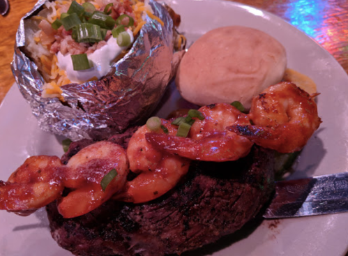A plate featuring a grilled steak topped with shrimp, a baked potato with toppings, and a dinner roll.