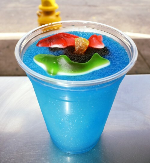 A clear cup filled with blue slushy ice, topped with gummy sharks and colorful candy.