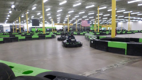 Go-karts racing on an indoor track with green and black barriers, featuring a large American flag in the background.