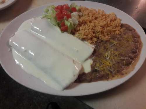 A plate of two enchiladas topped with white sauce, served with rice, refried beans, lettuce, and diced tomatoes.