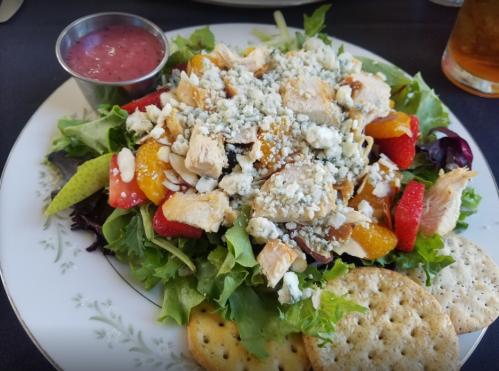 A colorful salad with greens, chicken, strawberries, mandarin oranges, blue cheese, and crackers, served with dressing.