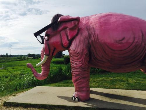 A large pink elephant statue wearing glasses stands on a concrete base against a green landscape and cloudy sky.