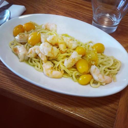A plate of pasta with shrimp and yellow tomatoes, served on a white dish with a glass of water in the background.
