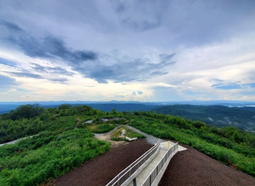 A scenic view from a mountain overlook, featuring lush greenery and a cloudy sky in the background.