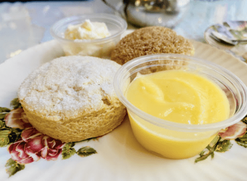 A plate with a sugar-dusted scone, a cookie, and a cup of lemon curd, garnished with floral designs.