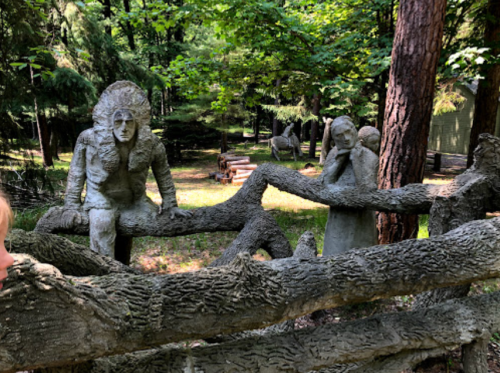 Sculptures of figures among trees, intertwined with a textured, natural-looking branch in a forest setting.