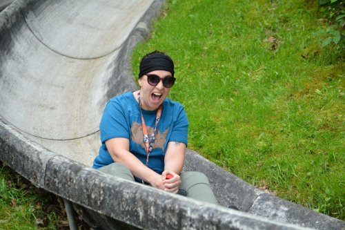 A person wearing sunglasses and a headscarf smiles excitedly while sitting in a slide surrounded by green grass.