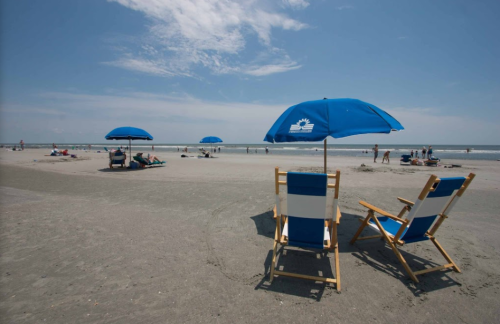 Two beach chairs under blue umbrellas on a sandy beach, with people enjoying the sun and ocean in the background.