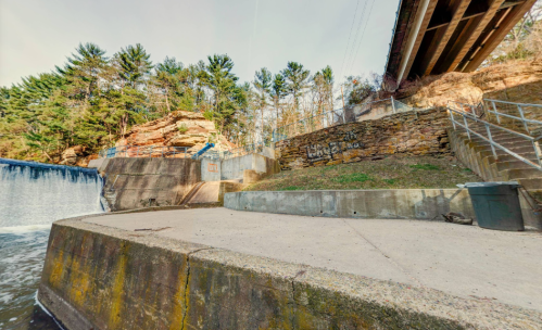 A concrete area by a waterfall, with a bridge overhead and rocky terrain lined with trees in the background.