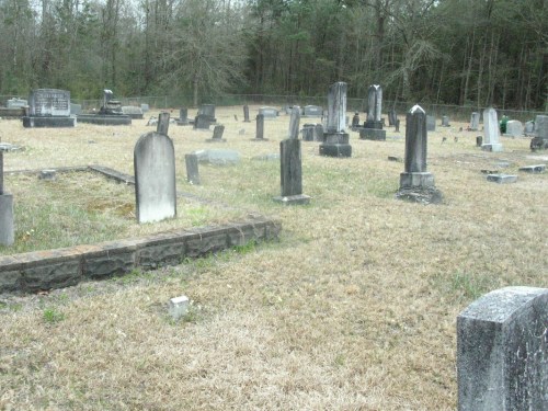 A cemetery with various gravestones and markers set in a grassy area surrounded by trees.