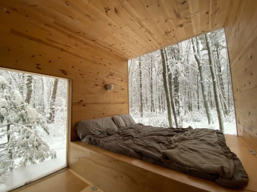 Cozy wooden cabin interior with a bed, surrounded by snowy trees outside a large window.