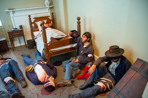 Civil War reenactors in a room, some lying on the floor, others seated, depicting a historical scene of battle aftermath.