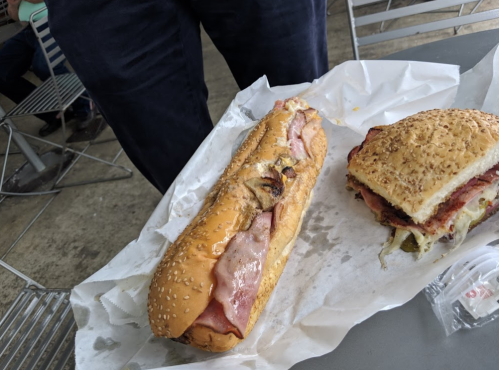 A sesame seed bun sandwich with ham and pickles, next to a smaller sandwich on a table.