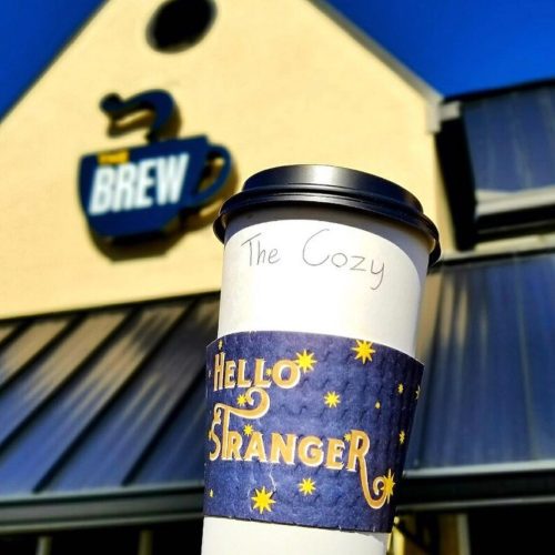 A coffee cup with a "Hello Stranger" sleeve in front of a café sign that reads "Brew." Clear blue sky above.