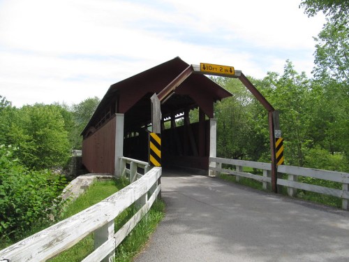 A wooden covered bridge with a sloped roof, surrounded by greenery and a gravel path leading to it.