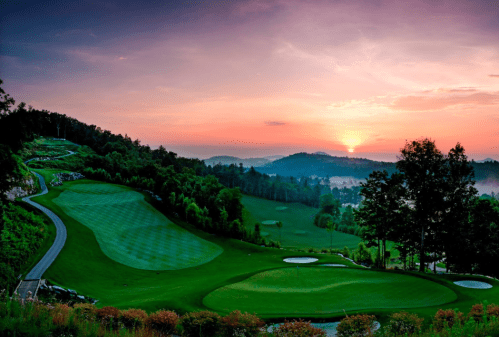 A scenic golf course at sunset, with lush green fairways and rolling hills under a colorful sky.