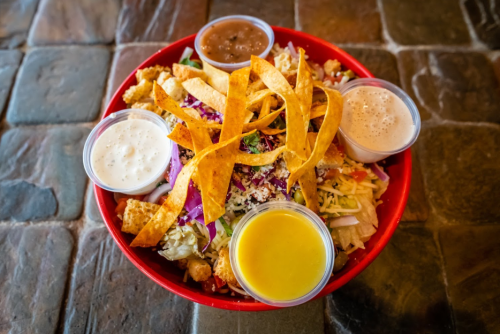 A colorful salad bowl topped with crispy tortilla strips and served with four small cups of dressing.