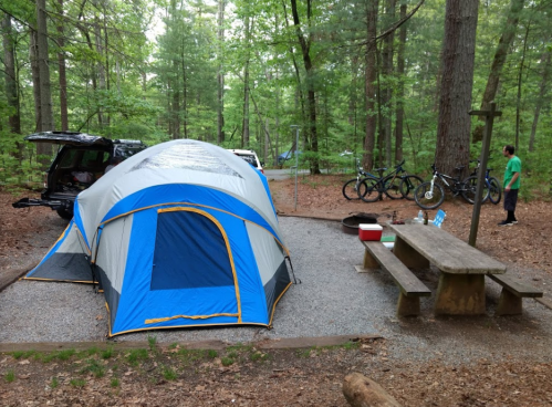 A blue and gray tent set up in a wooded campsite with a picnic table, fire pit, and bicycles nearby.