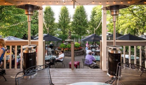 Outdoor dining area with tables, umbrellas, and greenery, featuring patio heaters and a statue in the center.