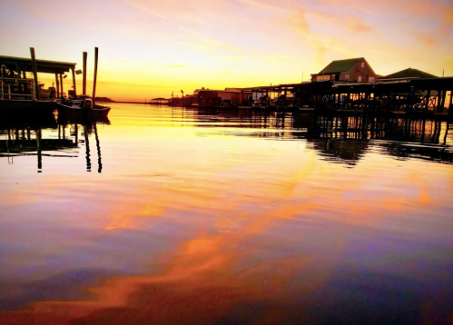 Sunset over calm water, reflecting vibrant colors and silhouettes of boats and piers along the shoreline.