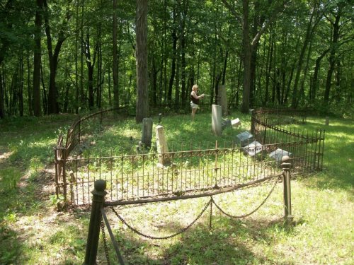 A person stands in a wooded cemetery, surrounded by old gravestones and a wrought iron fence.
