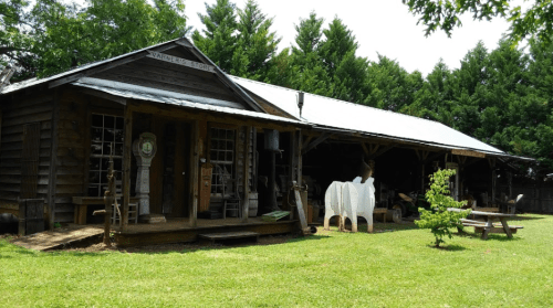A rustic wooden building with a porch, surrounded by greenery and outdoor seating, showcasing various vintage items.