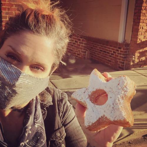 A person wearing a mask holds a star-shaped donut dusted with powdered sugar, smiling in sunlight.