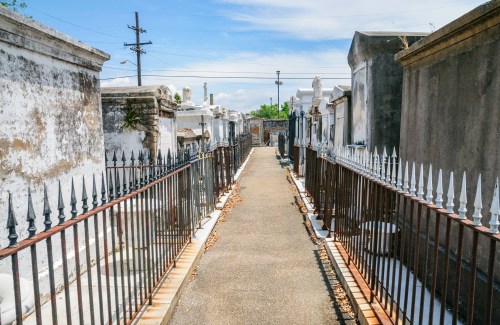 A narrow pathway lined with ornate iron fences in a cemetery, flanked by weathered tombs under a blue sky.
