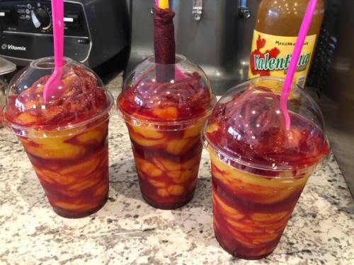 Three colorful cups filled with layered fruit, topped with a red sauce and a straw, sitting on a marble countertop.