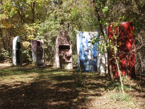 Five colorful, vintage car hoods stand upright in a wooded area, surrounded by trees and fallen leaves.