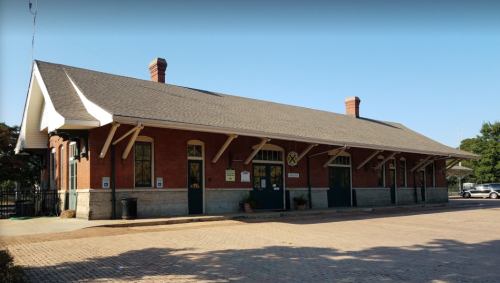Historic brick train station with a sloped roof and large windows, set against a clear blue sky.