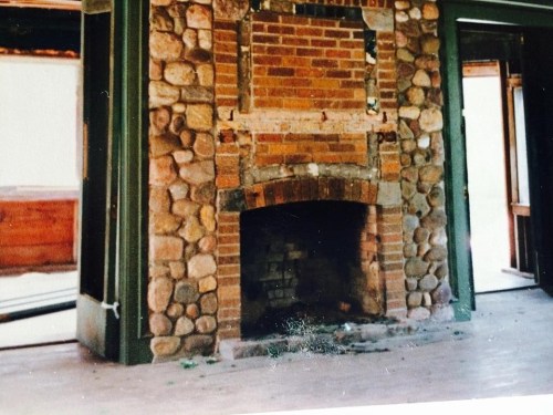 A rustic stone fireplace with a brick arch, set in an empty room with wooden floors and green walls.