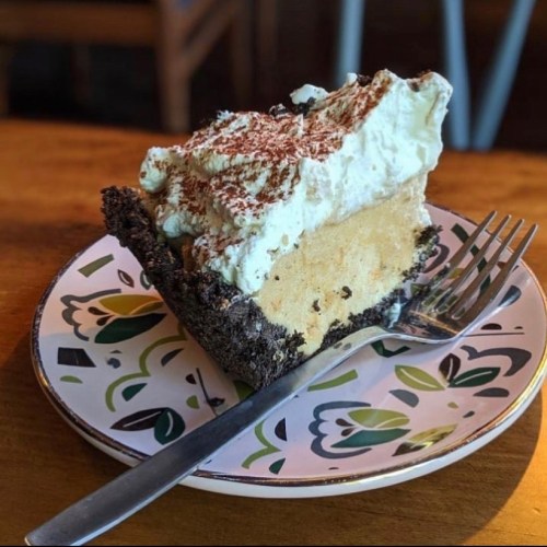 A slice of dessert pie with a chocolate crust, layered with creamy filling and topped with whipped cream and cocoa.