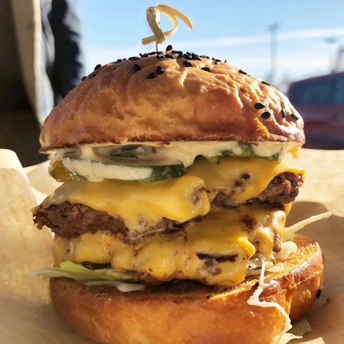 A towering cheeseburger with two beef patties, melted cheese, pickles, and lettuce on a toasted bun, topped with a skewer.