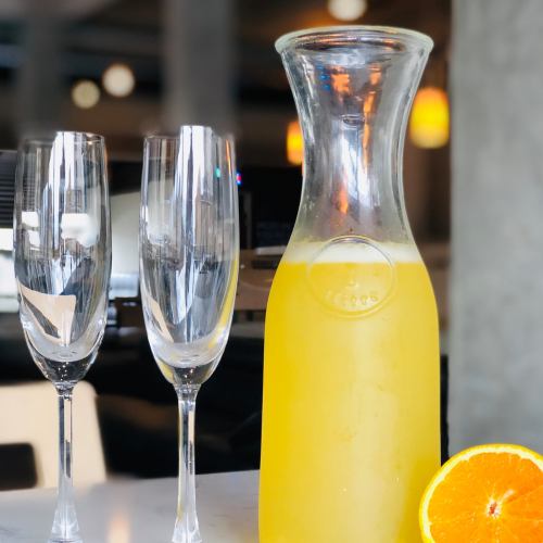 A glass pitcher of orange juice with two empty champagne flutes and a sliced orange on a table.